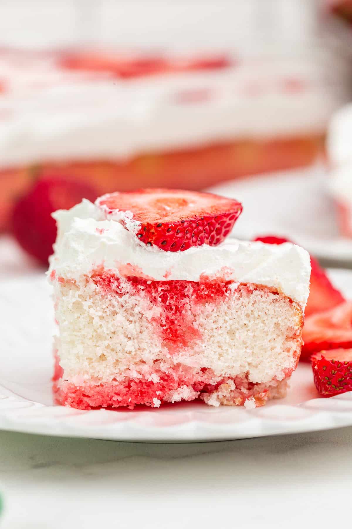 Slice of strawberry jello cake topped with whipped cream and fresh strawberries on a white plate.