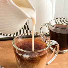 Clear mug of coffee with pitcher of coffee creamer being added to coffee.