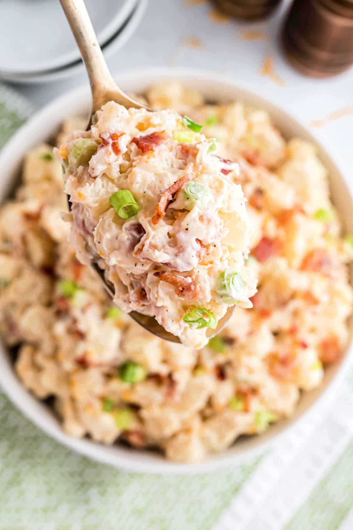 A spoonful of creamy loaded potato salad with bacon bits and green onions over a bowl of more potato salad.