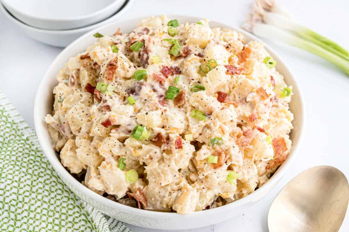 Creamy loaded potato salad with bacon bits and chopped green onions in a white bowl, beside a serving spoon.