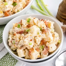 A bowl of loaded potato salad, topped with crispy bacon bits and chopped green onions, served with a spoon.