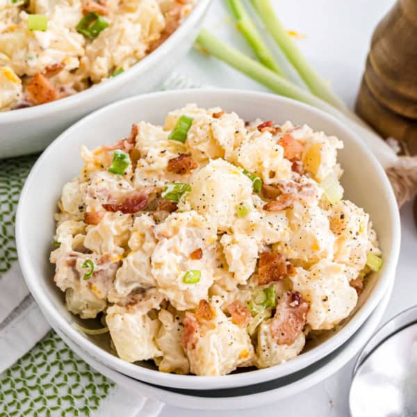 A bowl of loaded potato salad, topped with crispy bacon bits and chopped green onions, served with a spoon.