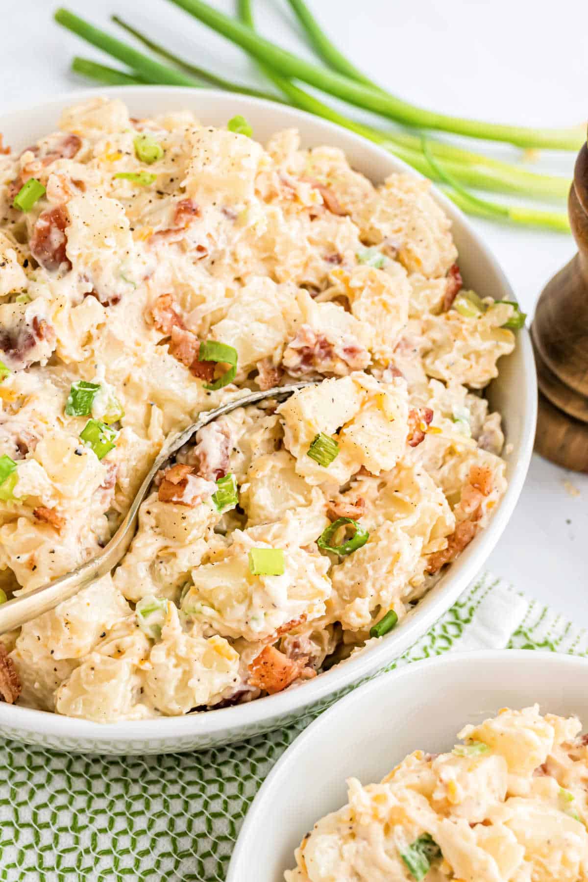 Bowl of creamy loaded potato salad with green onions and bacon, served with a spoon and fresh green onions nearby.