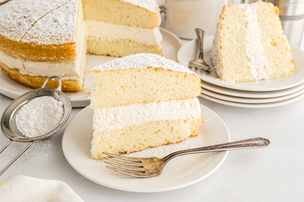 A slice of white cake with cream filling on a plate, made from a delicious Twinkie cake recipe, with more cake and plates in the background.