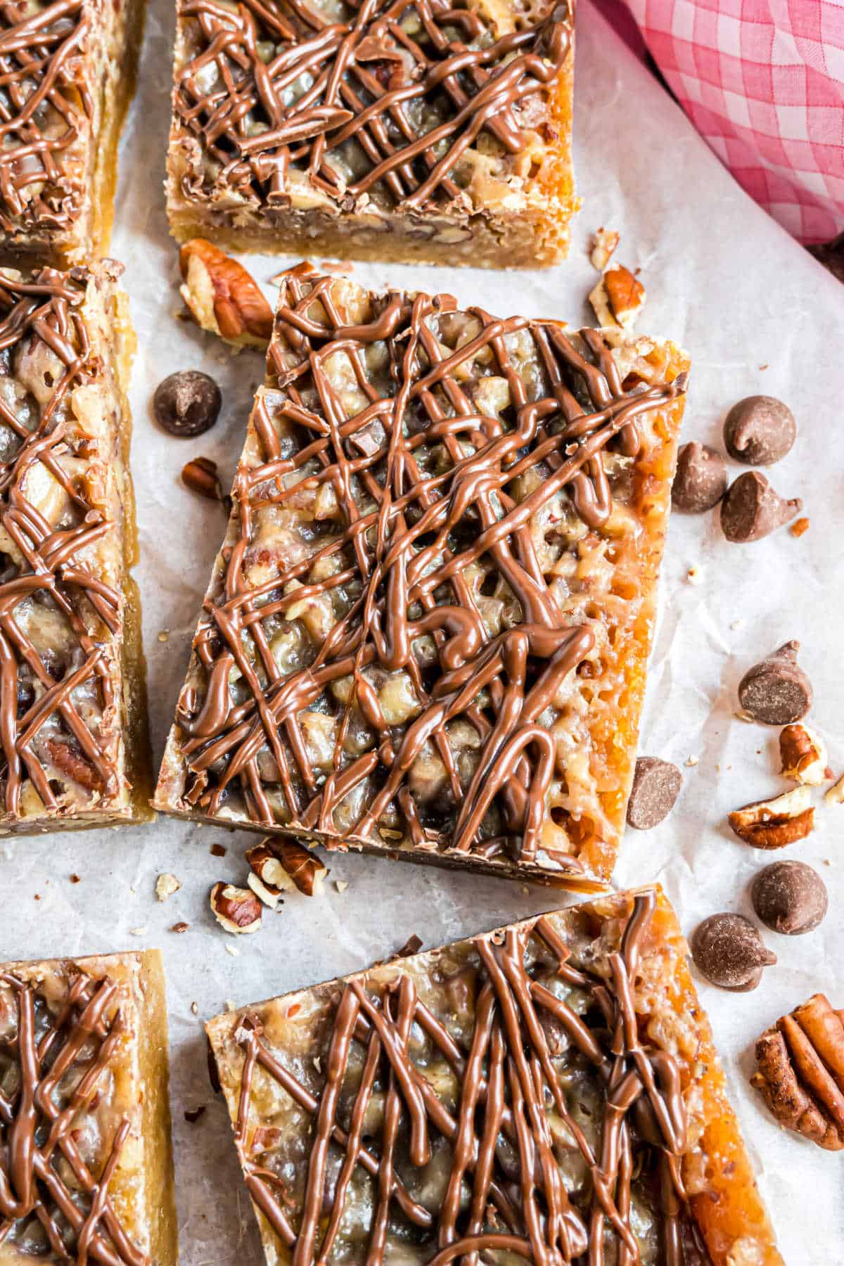 Square pecan pie blondies with chocolate drizzle on top, surrounded by chocolate chips and pecan pieces on parchment paper.