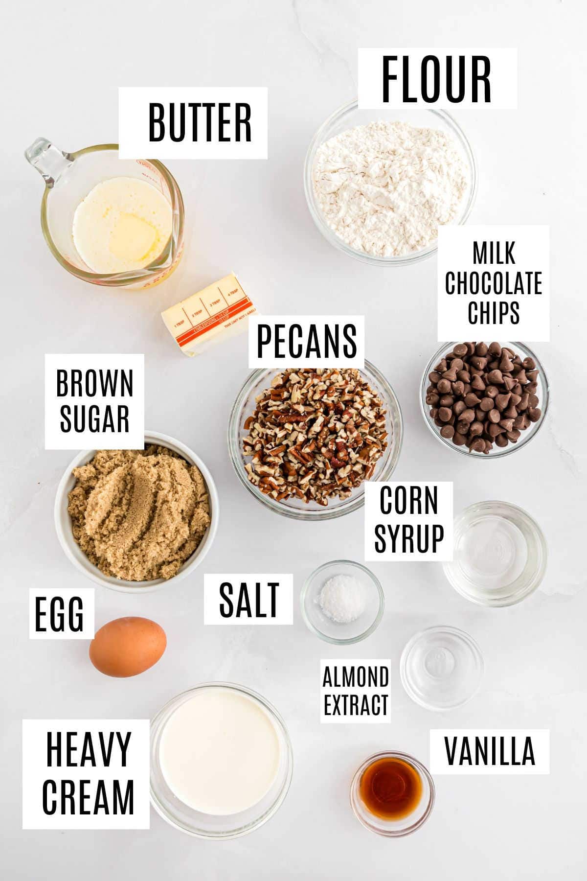 Baking ingredients labeled for delicious pecan pie blondies: butter, flour, brown sugar, pecans, milk chocolate chips, corn syrup, egg, salt, heavy cream, almond extract, and vanilla.