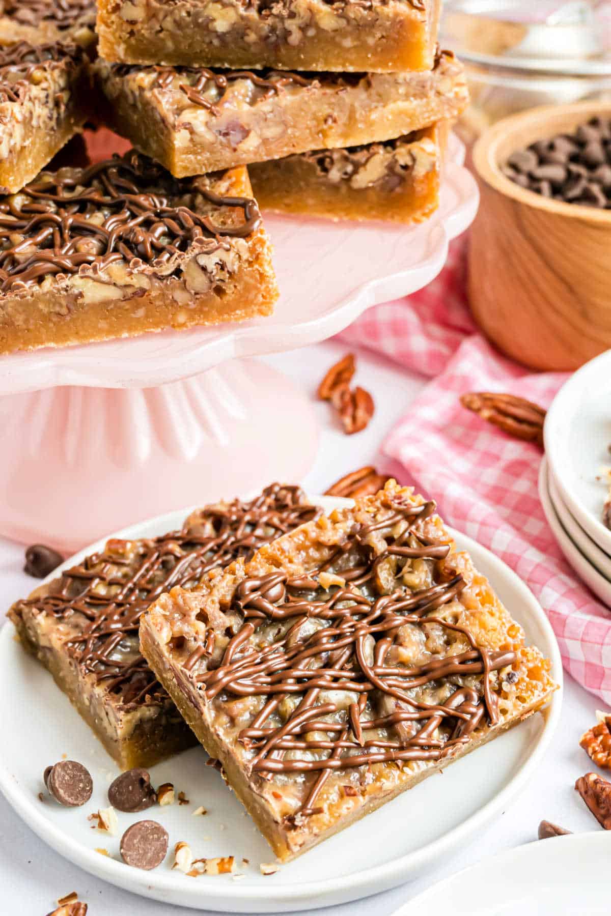 Pecan pie blondies drizzled with chocolate, stacked on a pink cake stand and white plate with chocolate chips nearby.