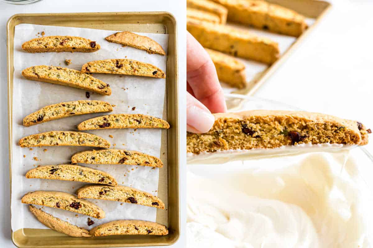 A hand holds a cranberry pistachio biscotti above a bowl of melted white chocolate, with more biscotti resting on the baking tray below.