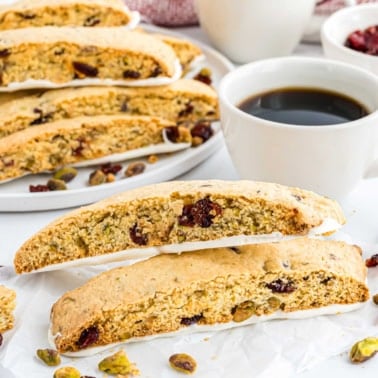 Two cranberry pistachio biscotti sit next to a cup of black coffee, with more tempting biscotti in the background.