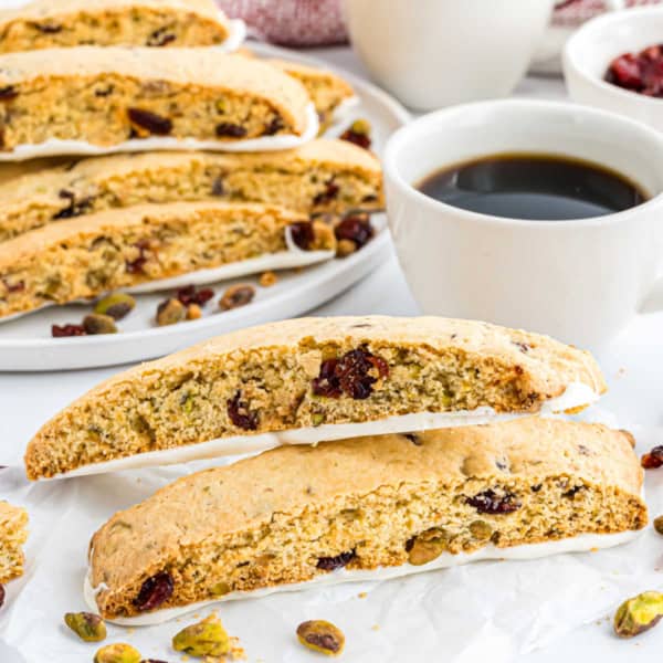 Two cranberry pistachio biscotti sit next to a cup of black coffee, with more tempting biscotti in the background.