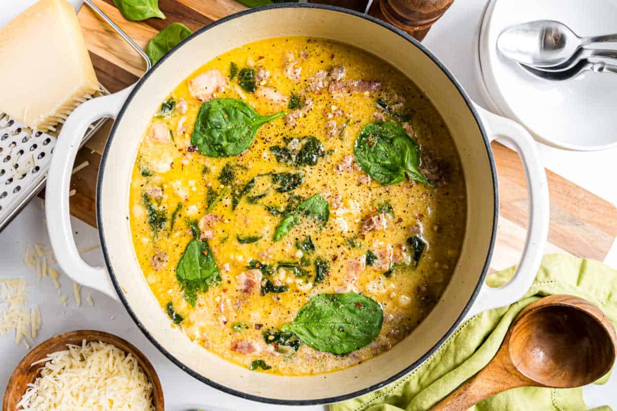 Creamy low carb keto zuppa toscana soup with spinach, bacon, and cheese in a white pot, surrounded by kitchen utensils and ingredients.