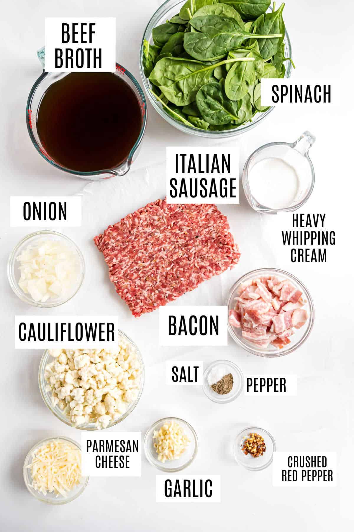 Labeled ingredients for a low carb keto Zuppa Toscana soup on a white surface, including sausage, spinach, broth, cream, bacon, and vegetables.