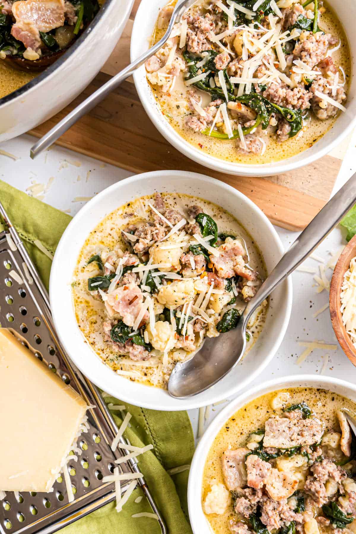 Three bowls of creamy low carb keto zuppa toscana with sausage, kale, and shredded cheese, served with a spoon and cheese grater nearby.