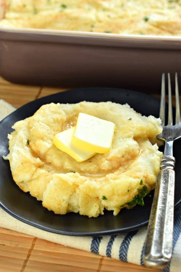 Plate of baked mashed potatoes with pat of butter, and fork. 13 x 9 dish of potatoes in background
