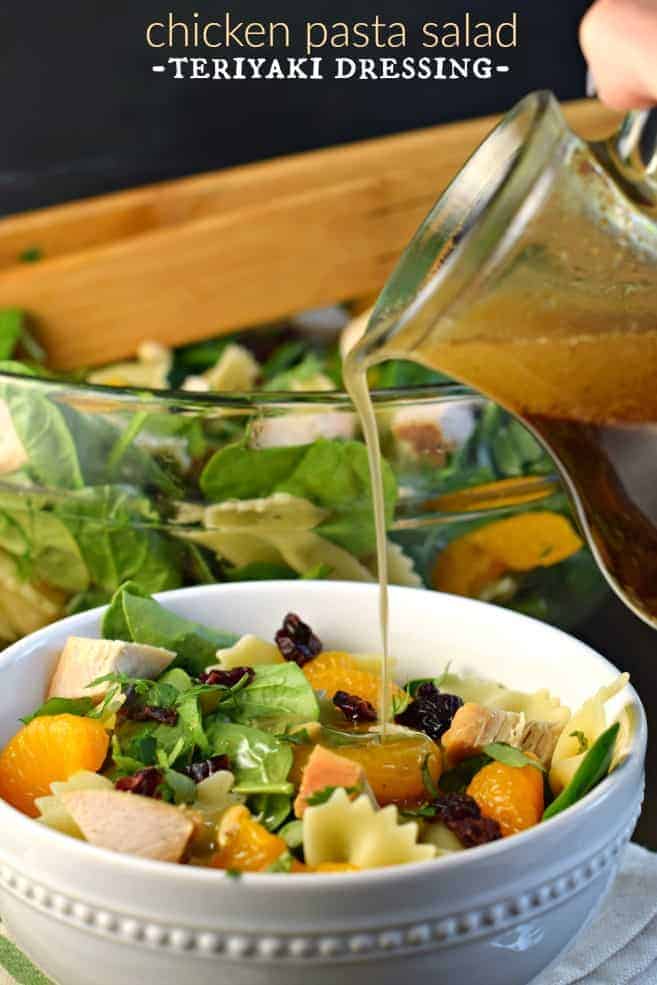 Chicken Pasta Salad with Teriyaki Dressing #mandarinoranges, #spinachsalad White salad bowl with spinach and pasta with chicken, mandarin oranges, and dressing being drizzled over the top.