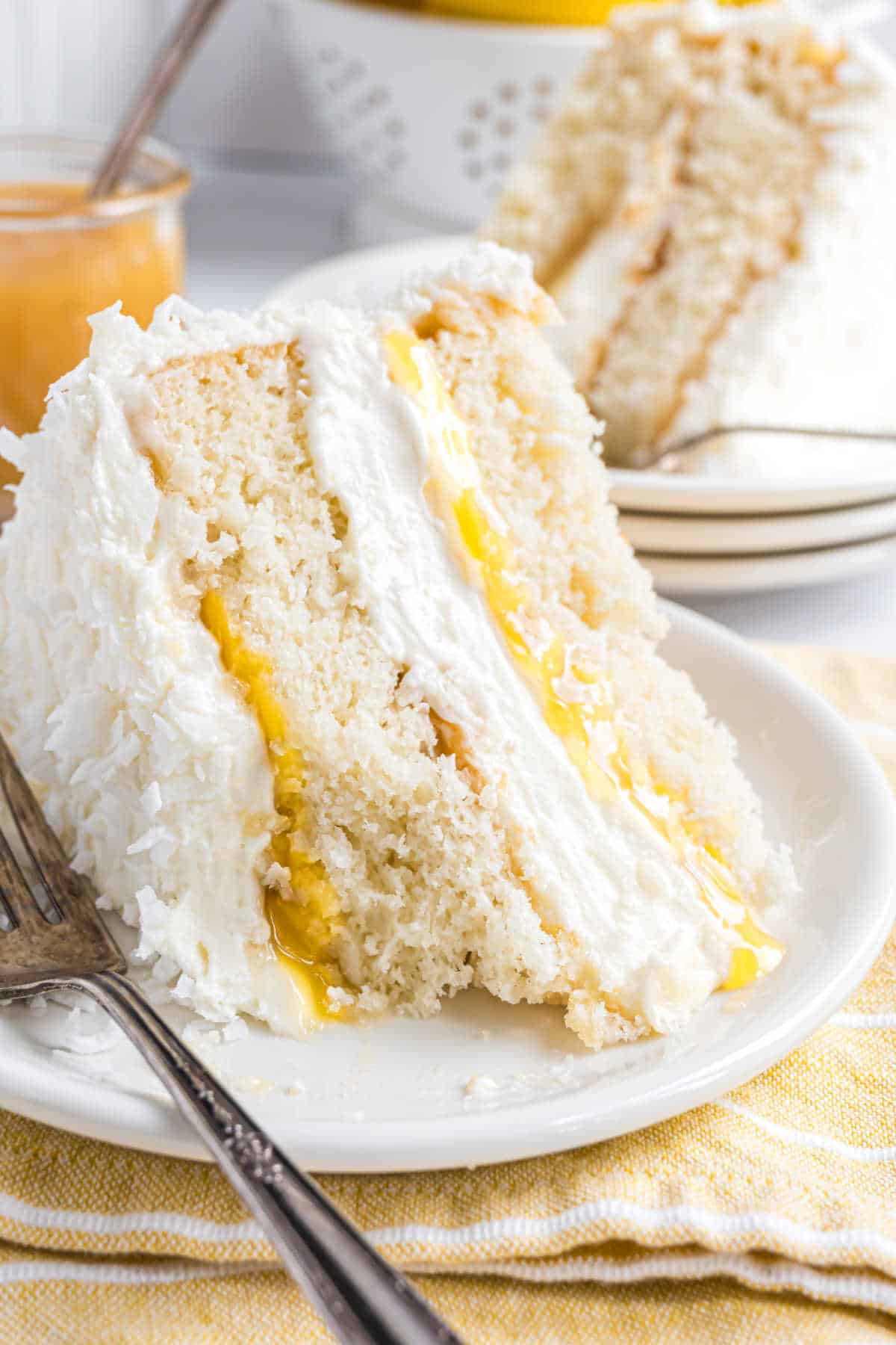 A slice of lemon coconut cake with creamy filling and yellow custard layers sits on a white plate next to a fork.