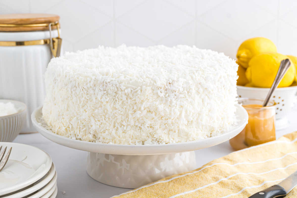 A white frosted lemon coconut cake sits on a cake stand, surrounded by fresh lemons and kitchen items.