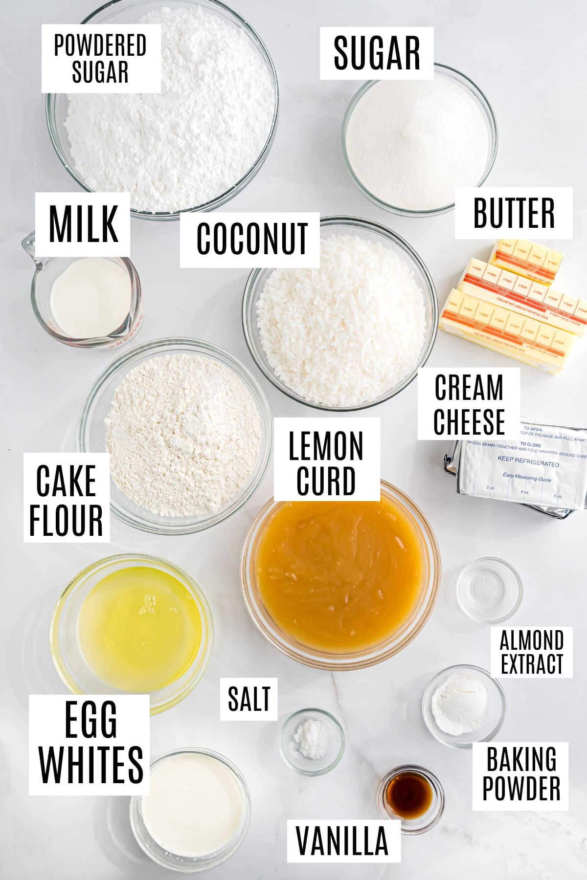 Various baking ingredients in bowls, each labeled: sugar, flour, milk, egg whites, coconut, butter, lemon curd—everything you need to create a delicious lemon coconut cake and more.