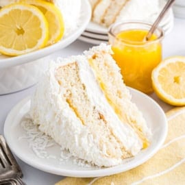 A slice of lemon coconut cake with lemon filling sits on a plate, garnished with coconut flakes and lemons nearby.