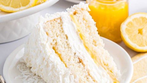 A slice of lemon coconut cake with lemon filling sits on a plate, garnished with coconut flakes and lemons nearby.