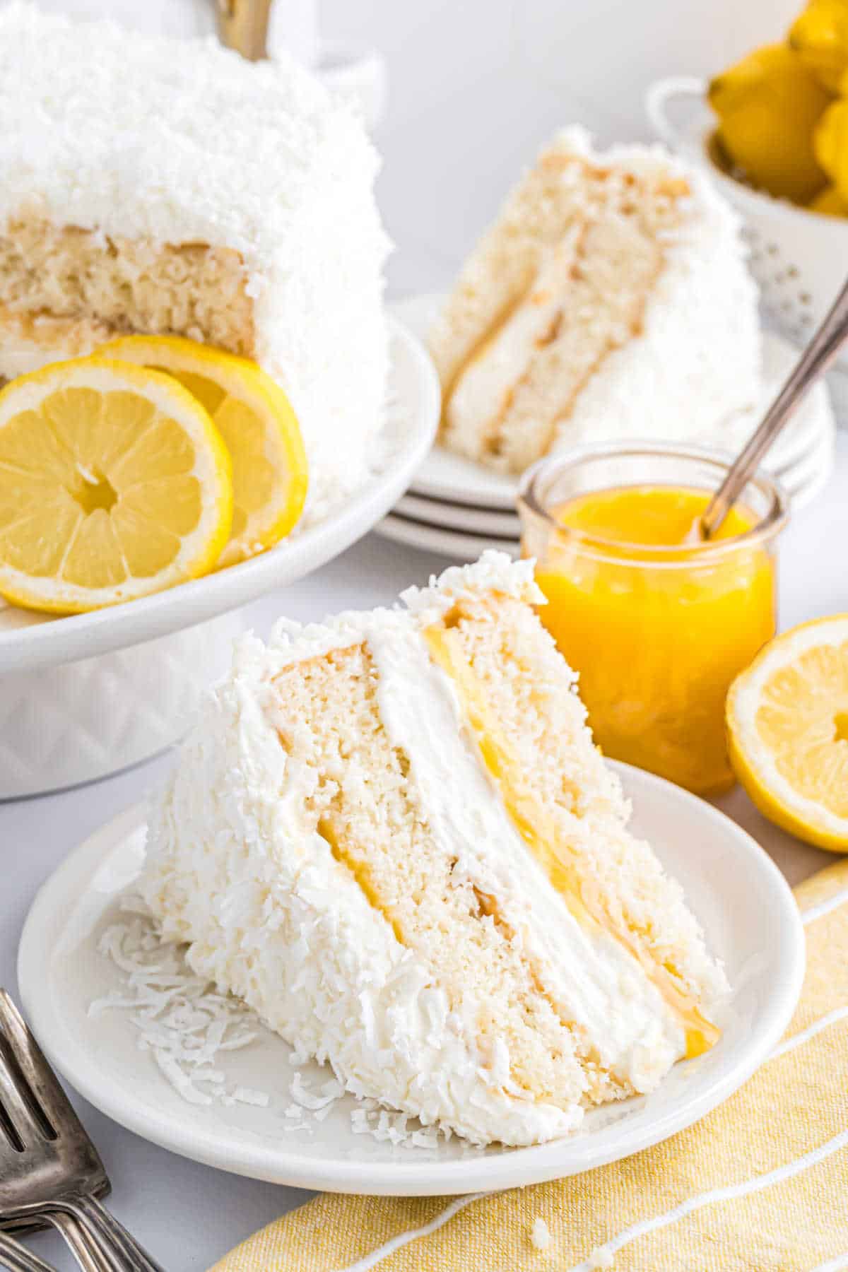 A slice of lemon coconut cake with lemon filling sits on a plate, surrounded by fresh lemon slices and a jar of tangy lemon curd.