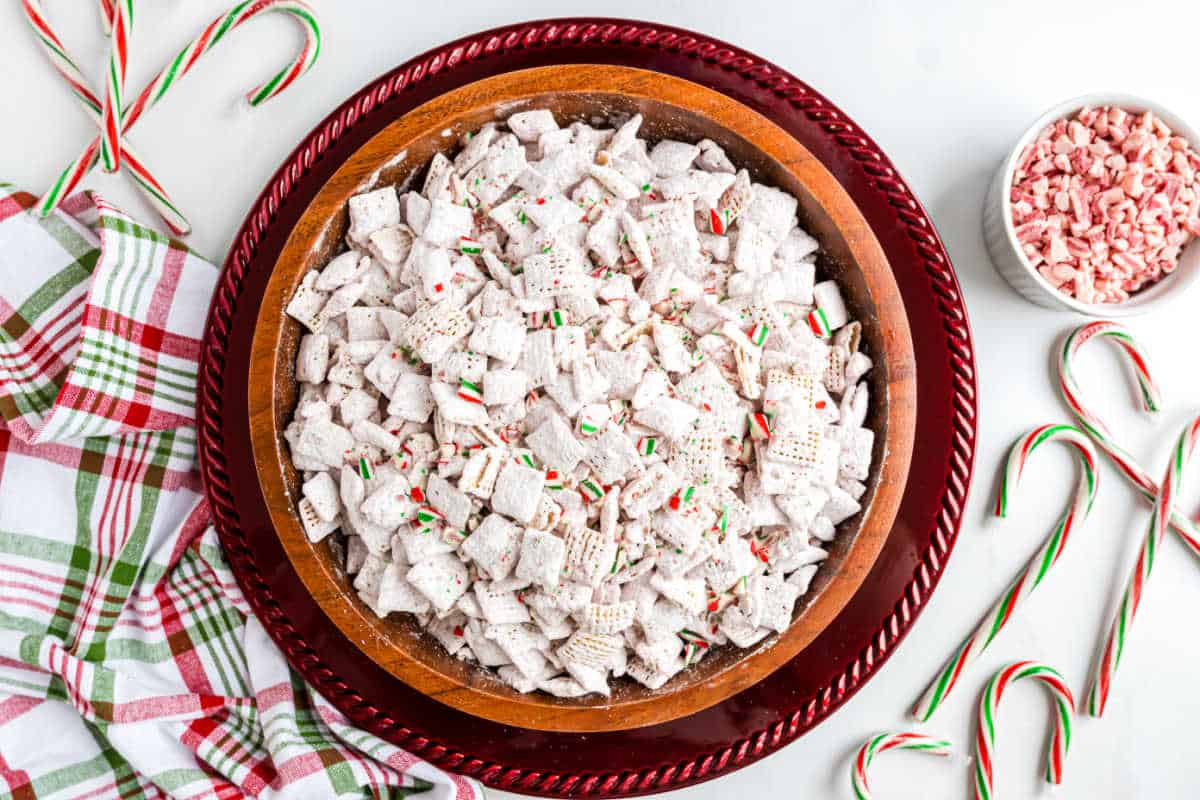 Large bowl of peppermint puppy chow Chex mix dusted with powdered sugar, surrounded by candy canes and a festive holiday napkin.