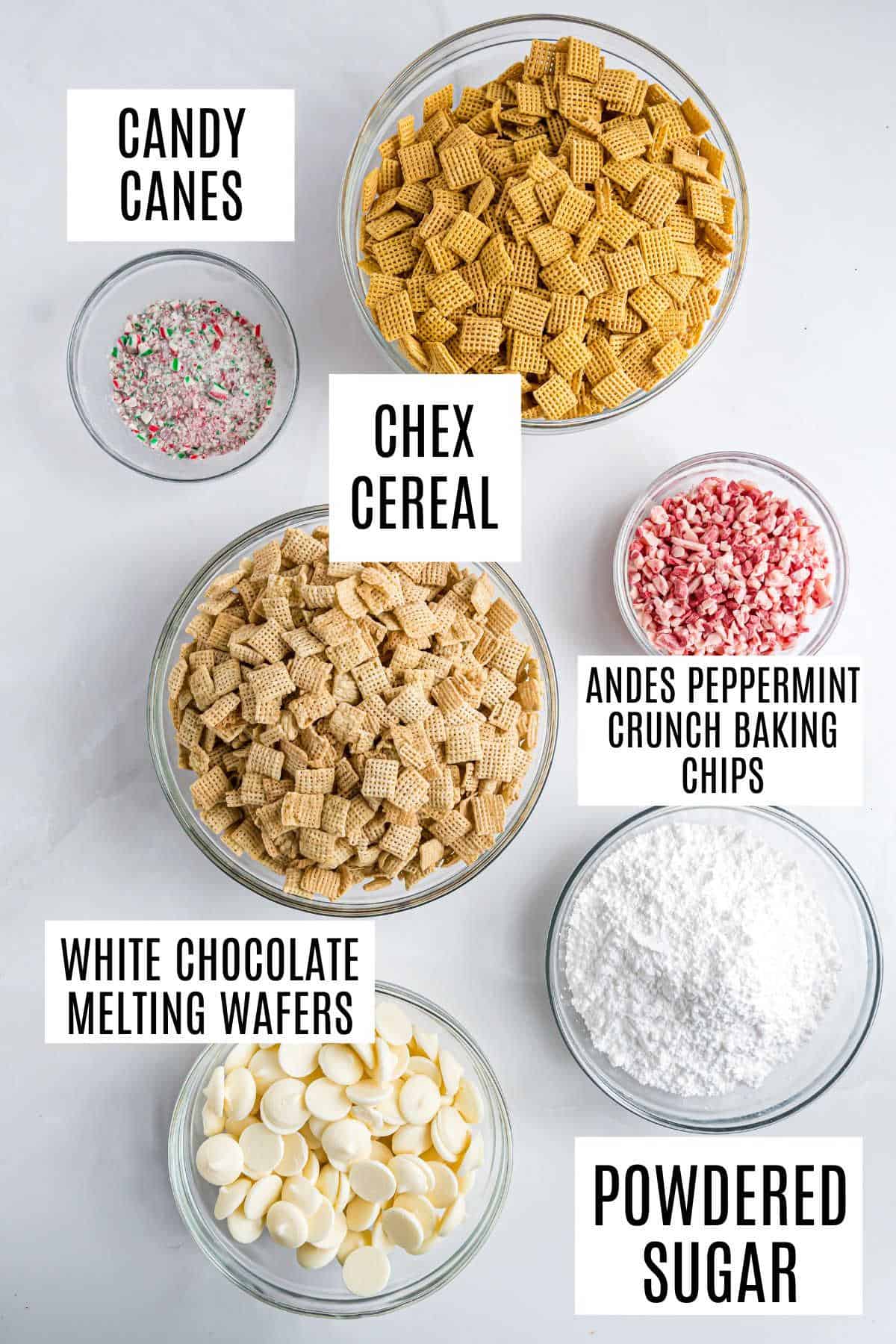Bowls of Chex cereal, white chocolate wafers, Andes chips, crushed candy canes, and powdered sugar on a white surface—perfect ingredients for making festive peppermint puppy chow.