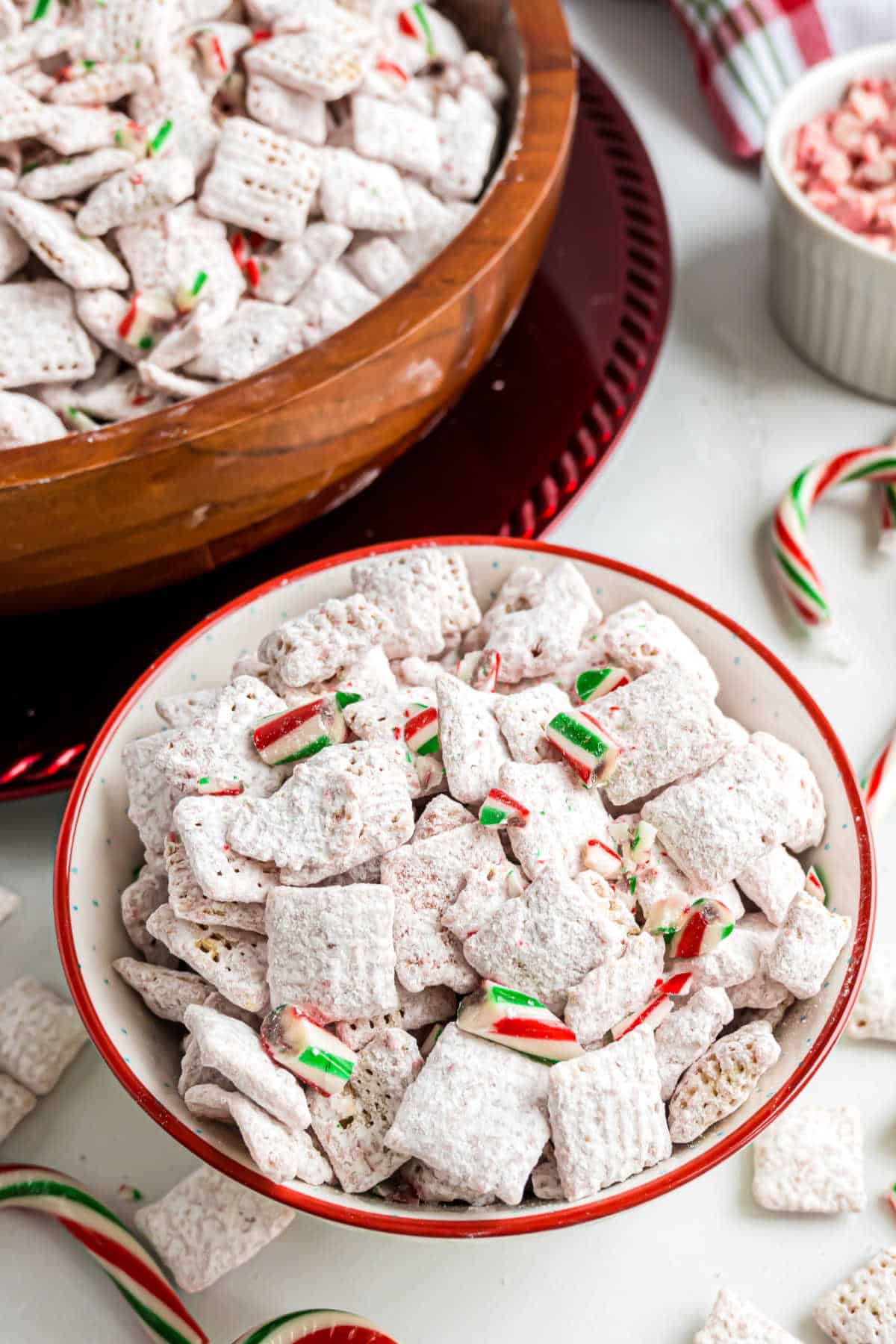 A bowl of peppermint puppy chow snack mix with crushed candy canes sits beside a larger bowl and whole candy canes, ready for a festive treat.