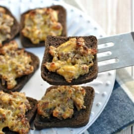 Rye bread toasts topped with a cheesy sausage mixture—just like the classic hanky panky recipe—rest on a white plate, one being lifted with a spatula.