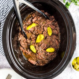Slow cooker with mississippi pot roast shredded and set of tongs to serve.