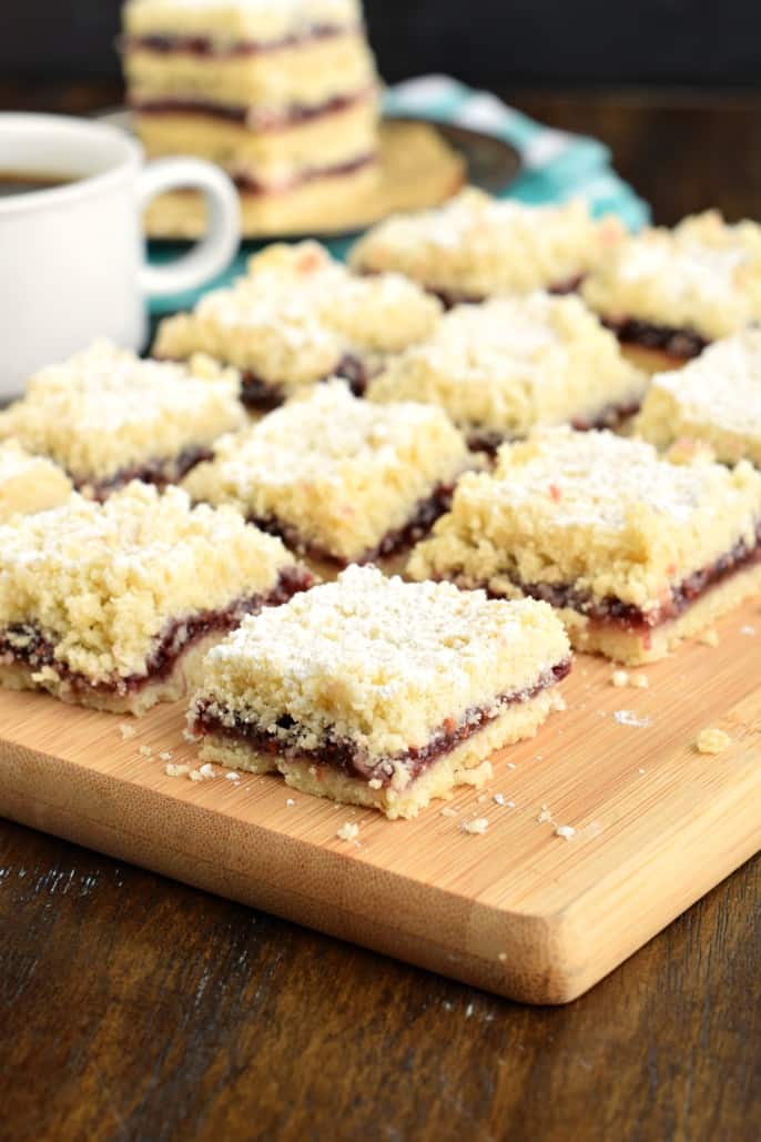 Shortbread cookie bars with a raspberry filling and powdered sugar on top, cut into squares on a wooden cutting board.