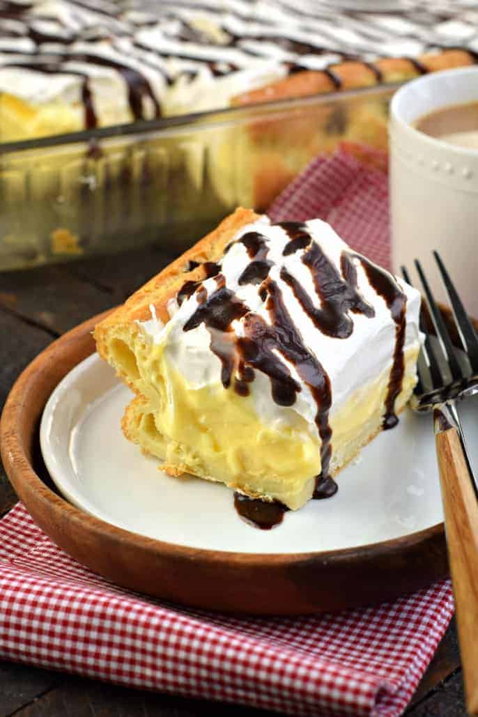 Big slice of cream puff cake on a white plate and wooden charger. Served on a red checkered napkin with a white mug of coffee.