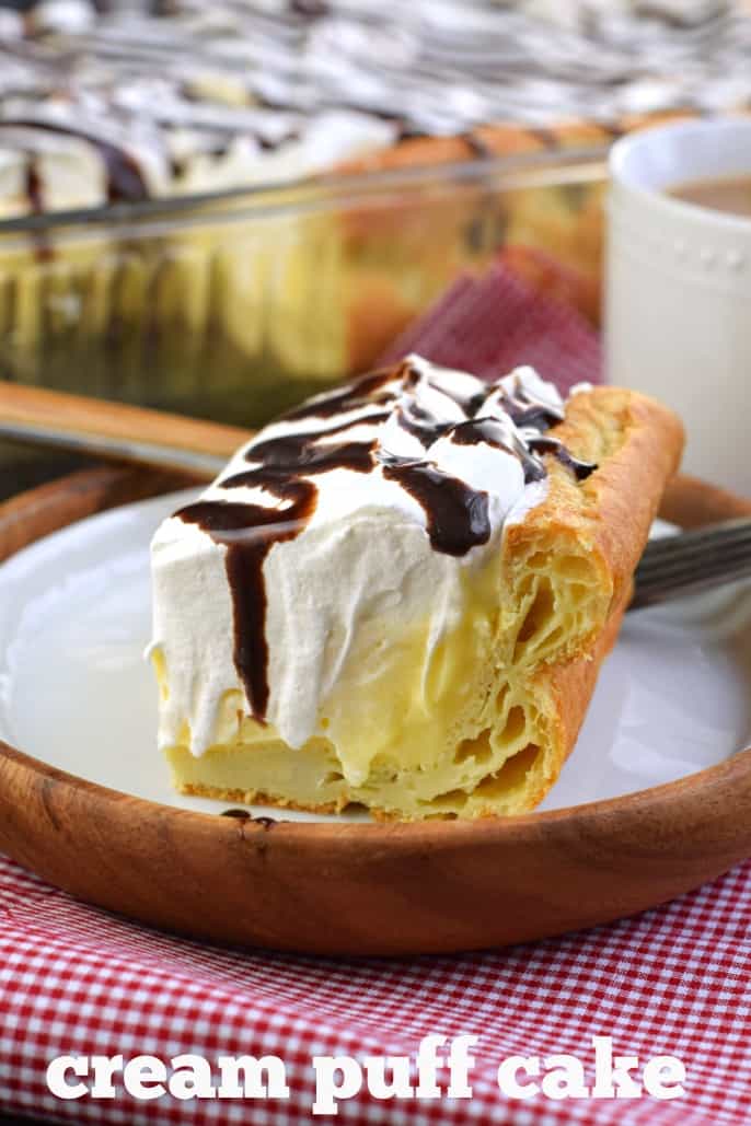 Slice of cream puff cake with flaky crust and topped with chocolate syrup, on a wooden plate and red checkered napkin.