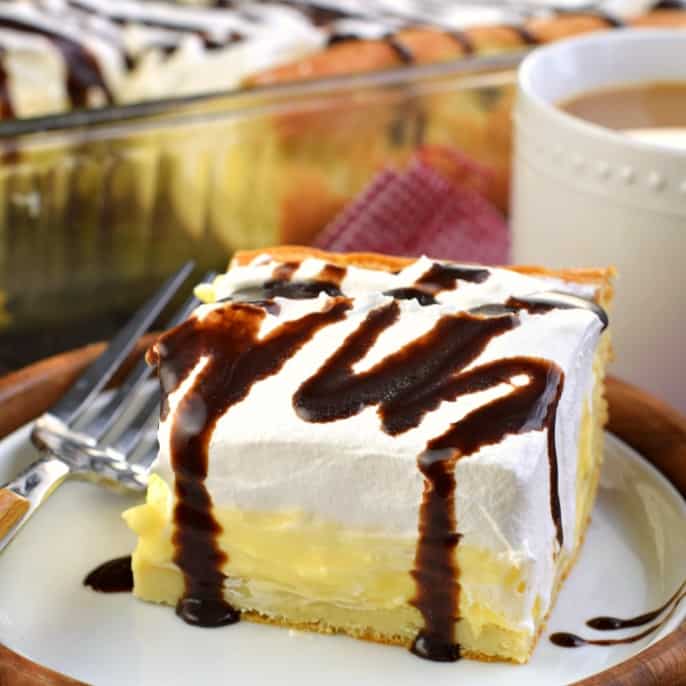 Slice of cream puff cake topped with chocolate syrup and cup of coffee in background.