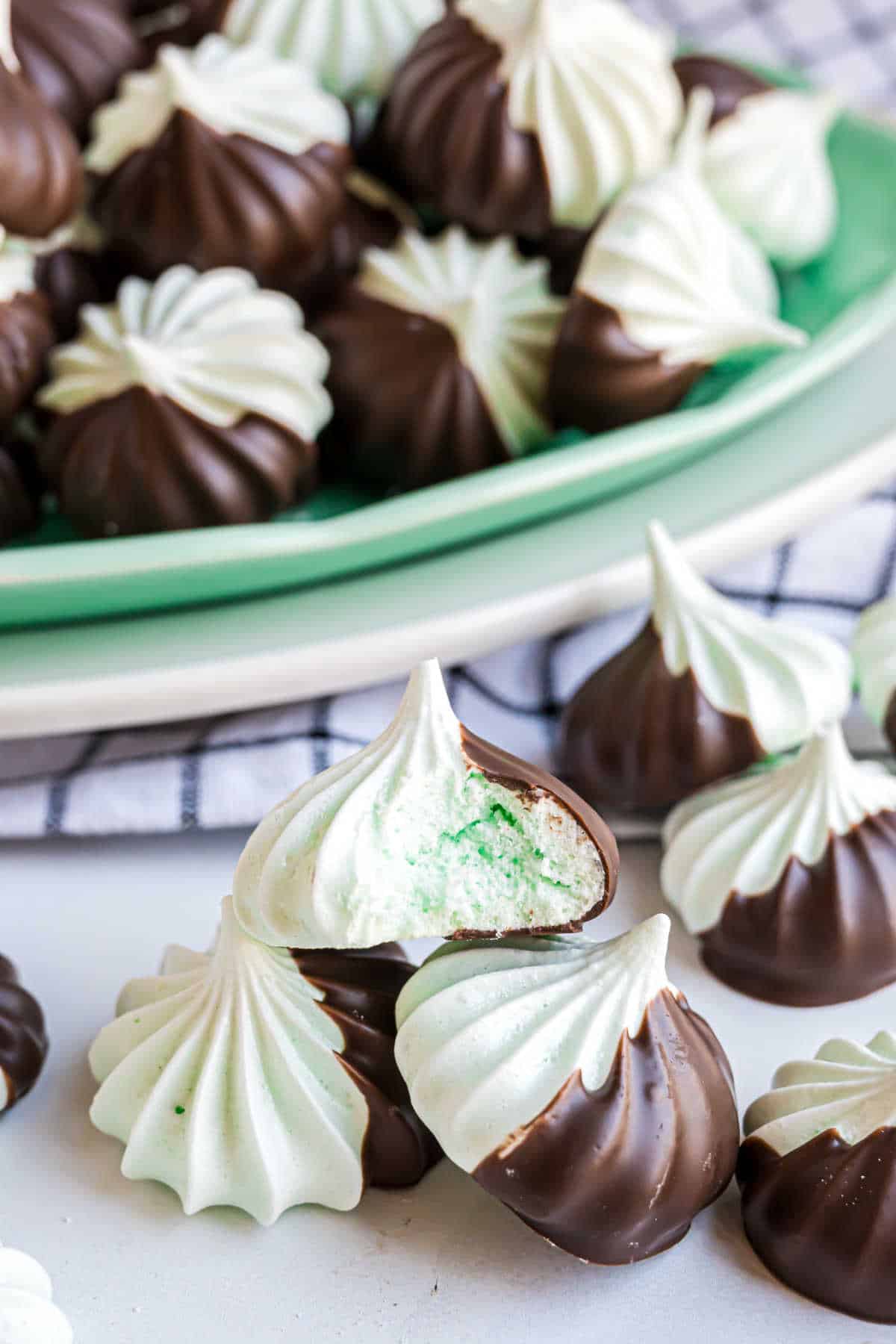 Chocolate mint meringues dipped in chocolate are stacked on a plate, with one cookie broken open to reveal its airy center.