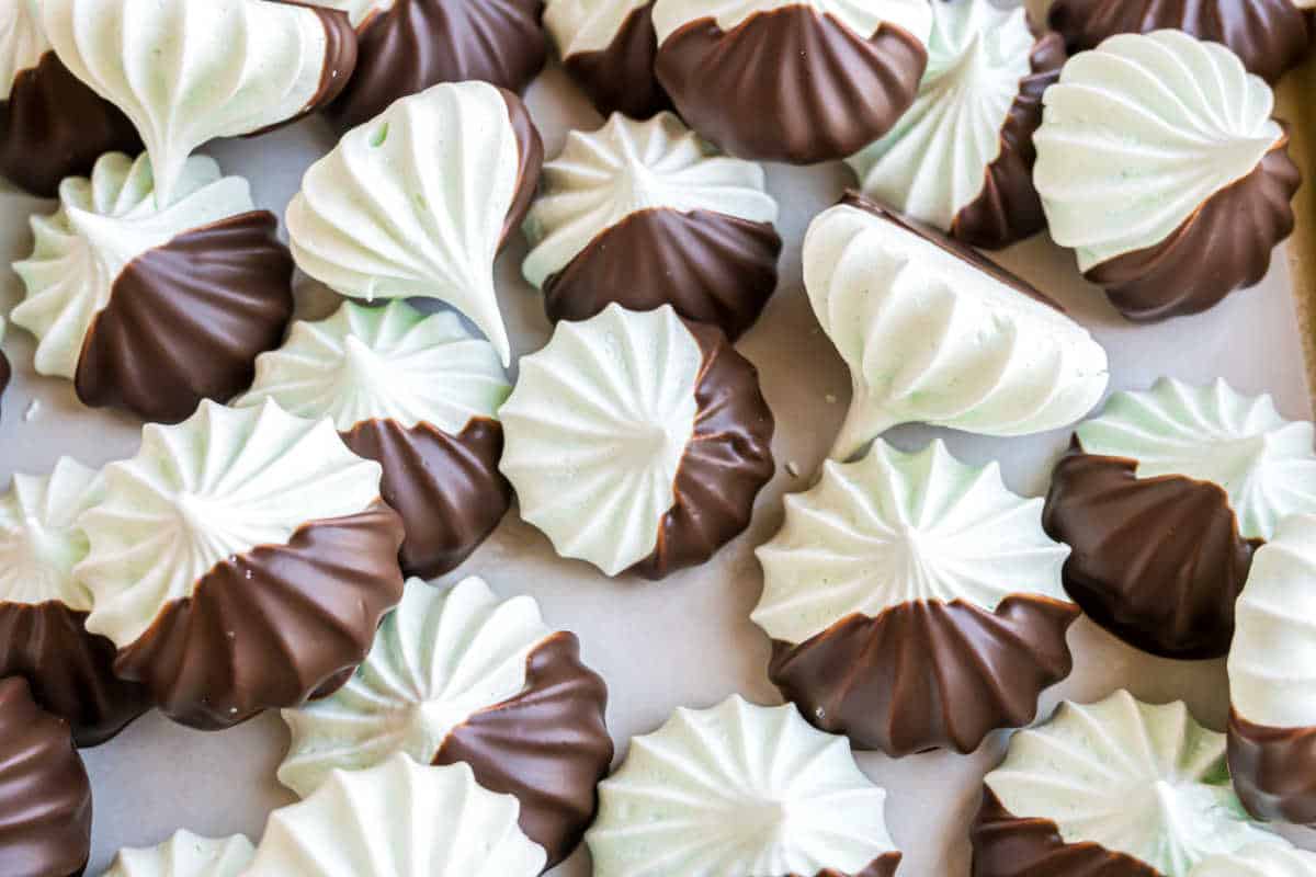 Chocolate mint meringues in a refreshing mint-green hue, half-dipped in chocolate, are arranged closely on a light surface.