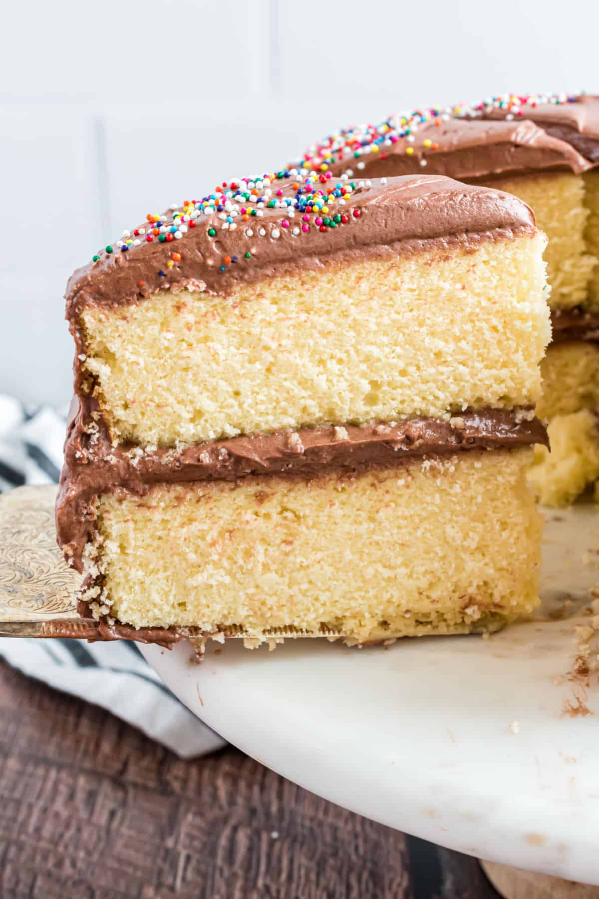 Two layers of yellow cake with chocolate frosting and sprinkles on a cake platter.