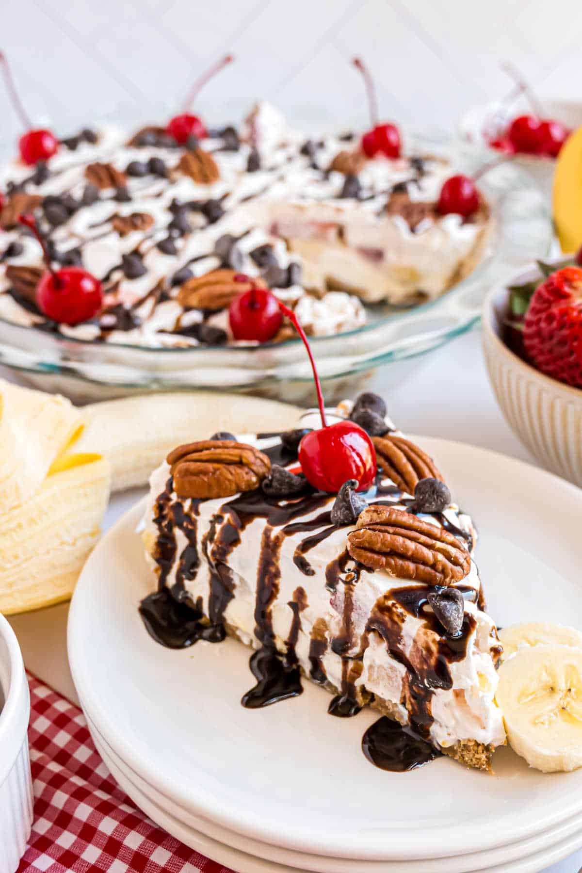 Slice of banana split cheesecake with chocolate syrup on a white dessert plate.