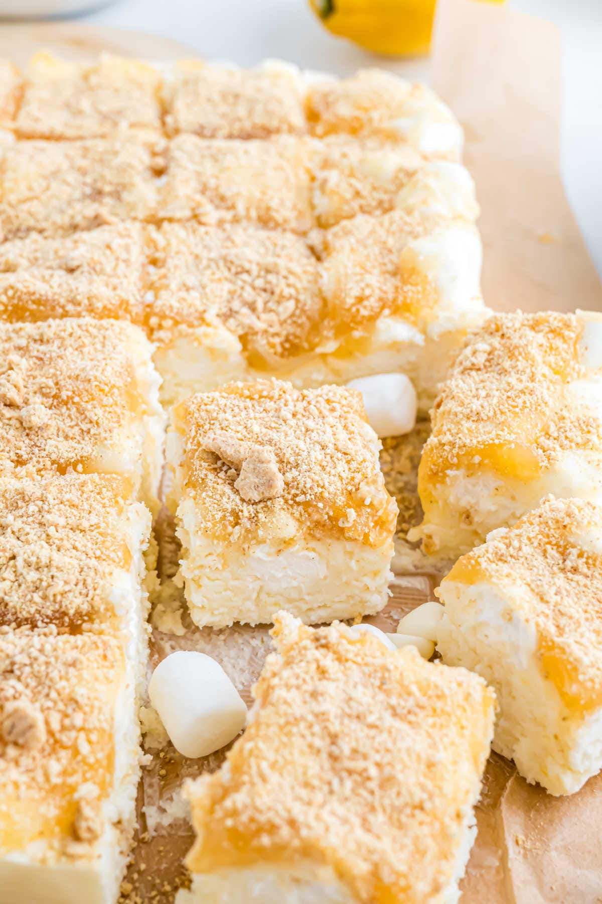 Lemon cheesecake bars topped with crumbs, cut into squares, and surrounded by mini marshmallows for a playful touch—perfect for fans of lemon meringue fudge.