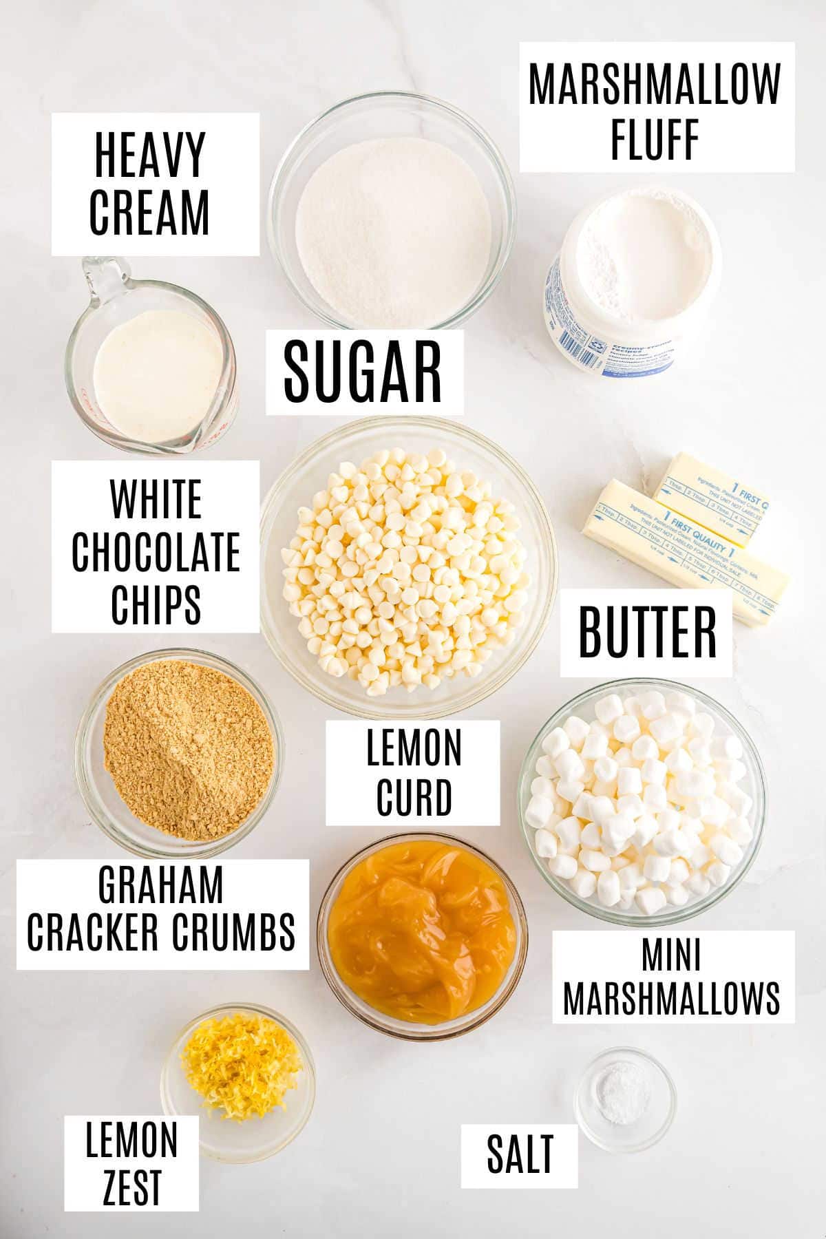 Labeled baking ingredients in bowls: heavy cream, sugar, marshmallow fluff, white chocolate chips, butter, graham cracker crumbs, lemon curd, mini marshmallows, lemon zest, and salt—perfect for making lemon meringue fudge.