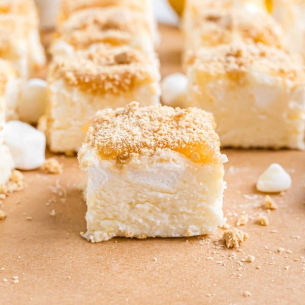 A close-up of creamy lemon meringue fudge dessert bars topped with crumbs, surrounded by mini marshmallows and white chocolate chips.