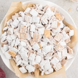 A bowl of snickerdoodle muddy buddies—a powdered sugar-coated cereal snack mix—rests on parchment paper.