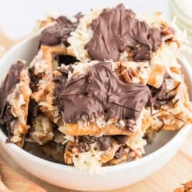 A bowl filled with caramel coconut cracker toffee, nut, and chocolate-topped dessert bars arranged in a stack.