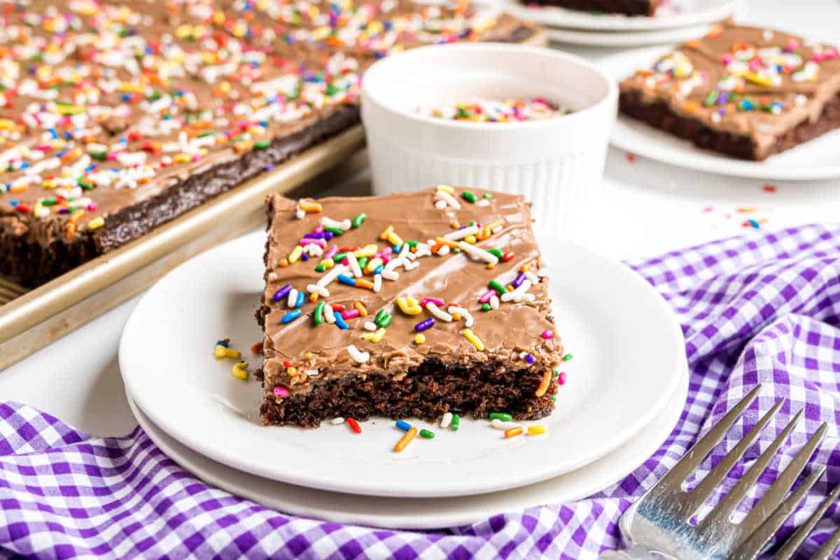 A slice of chocolate buttermilk sheet cake with frosting and rainbow sprinkles sits on a white plate beside a fork.