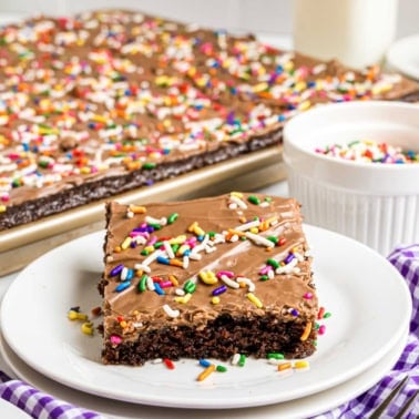 A frosted chocolate buttermilk sheet cake slice with colorful sprinkles sits on a plate, with more cake and sprinkles in the background.