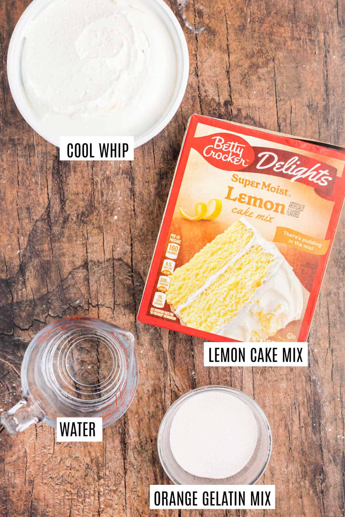 Top view of ingredients for a lemon orange Jell-O cake: Cool Whip, lemon cake mix, orange gelatin mix, and a cup of water on a wooden surface.