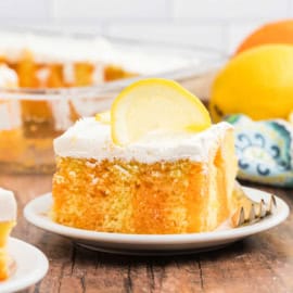 A slice of lemon orange jell-o cake with white frosting and a lemon slice on top, served on a white plate with a fork.