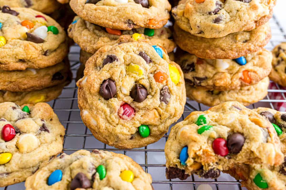 Colorful pudding cookies studded with chocolate chips and candy pieces cooling on a wire rack, one with a bite taken out.