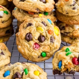 Chocolate chip and candy pudding cookies stacked on a cooling rack, with one cookie in focus and one with a bite taken.