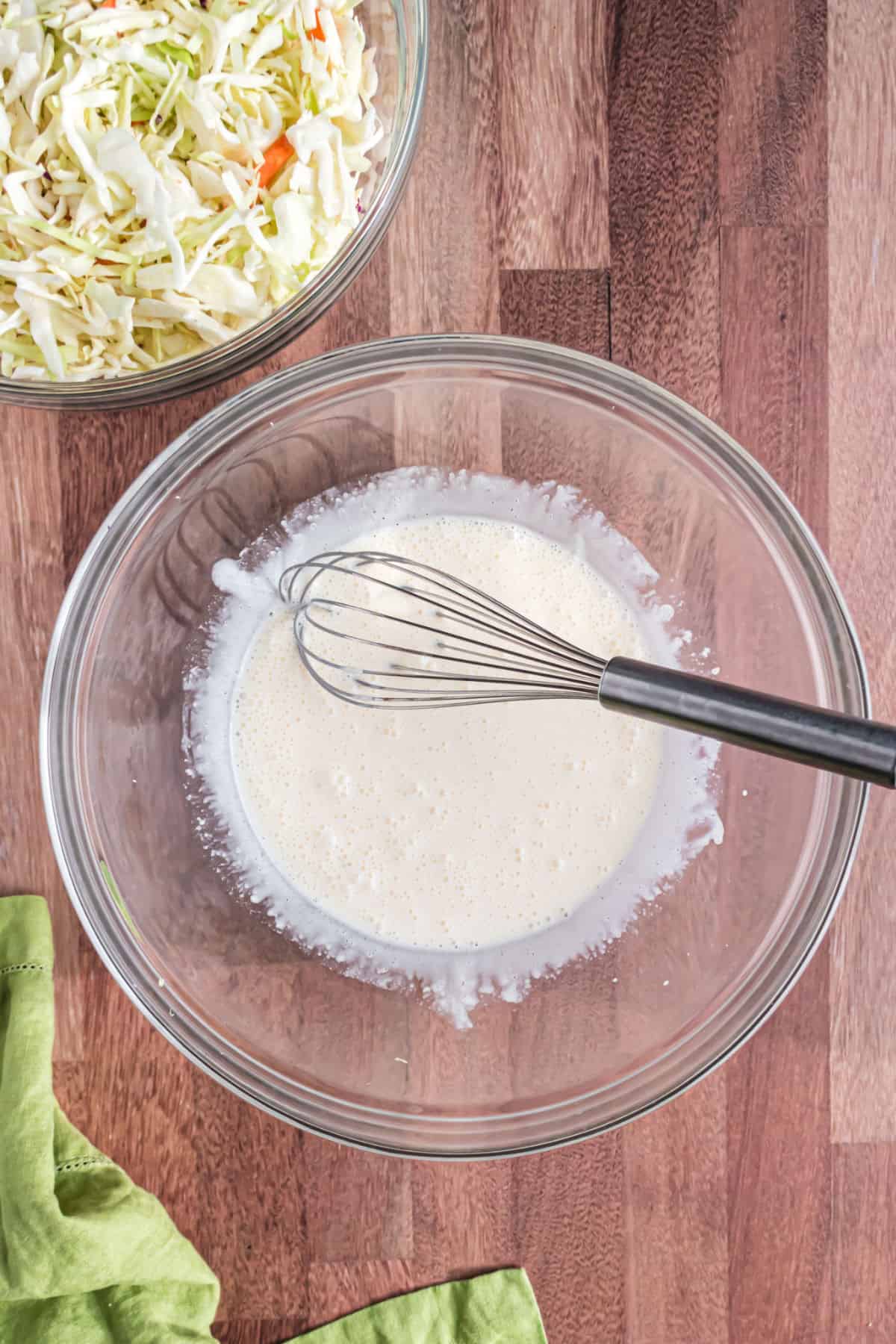 Dressing for coleslaw in a clear glass bowl with a whisk.